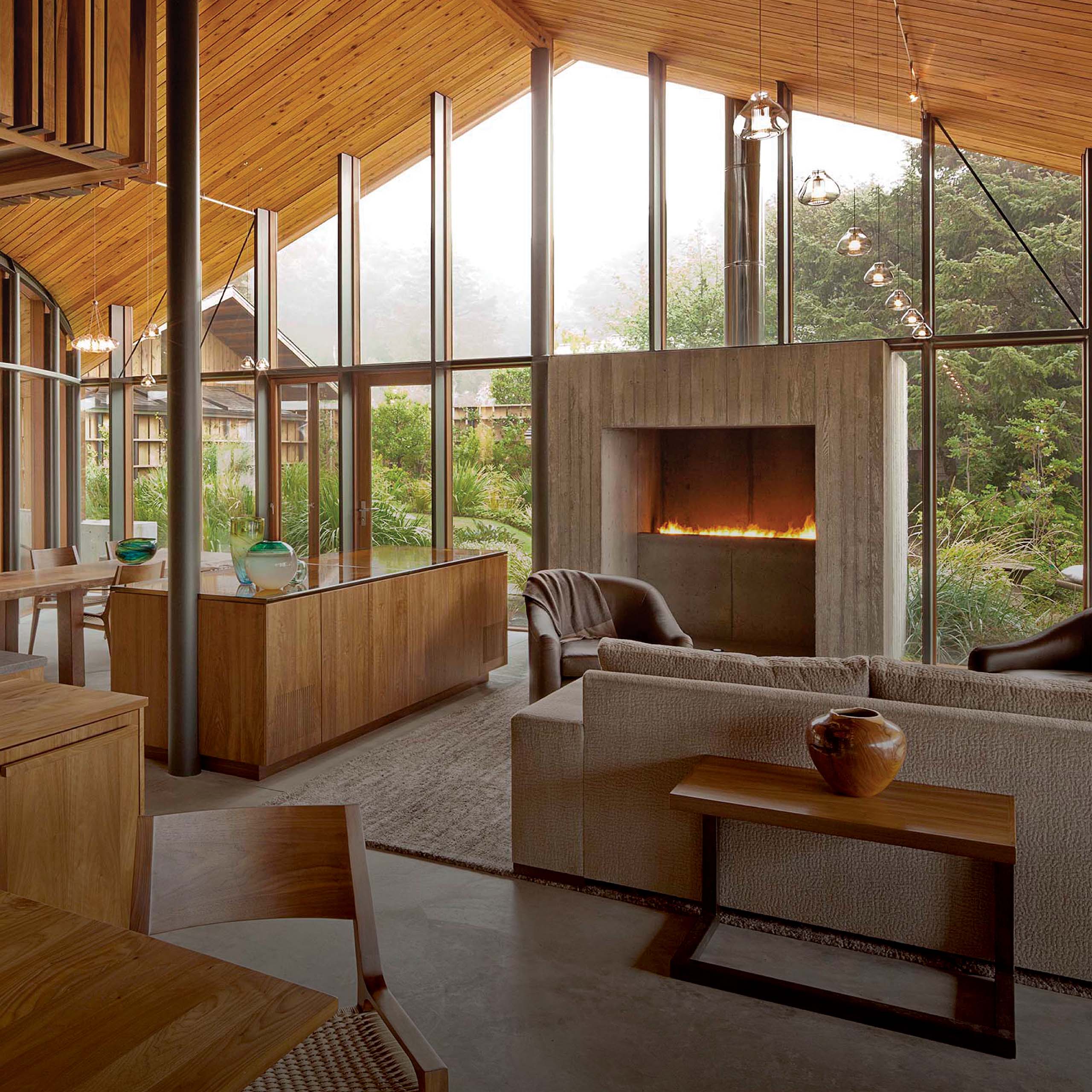 Interior of Singing Sands Cottage by Jones Studio Inc showing cedar ceiling, floor-to-ceiling glass walls, board-formed concrete fireplace, and views of coastal garden — designed by Eddie Jones FAIA