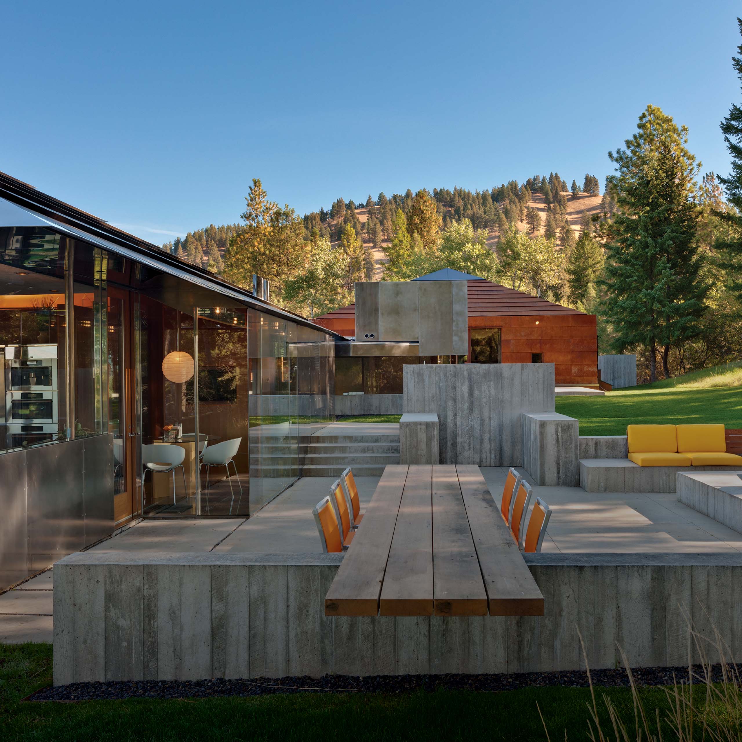 Outdoor dining patio at Starwood Residence by Eddie Jones FAIA Jones Studio Inc Missoula Montana showing board-formed concrete dining table wood benches orange chairs yellow outdoor seating glass kitchen pavilion Cor-Ten steel residence and Montana mountain ridge