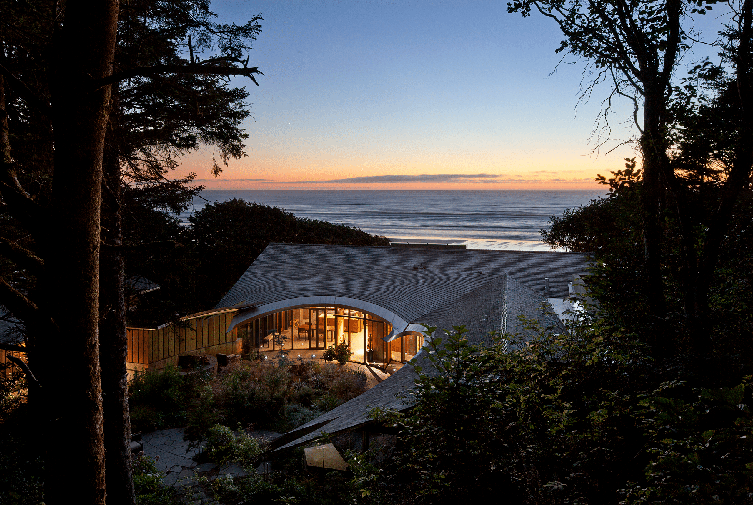 ARA – Singing Sands Cottage Exterior by Eddie Jones Singing Sands Cottage at dusk by architect Eddie Jones FAIA of Jones Studio Inc, Cannon Beach Oregon, showing curved cedar and slate roof with glowing interior and Pacific Ocean view